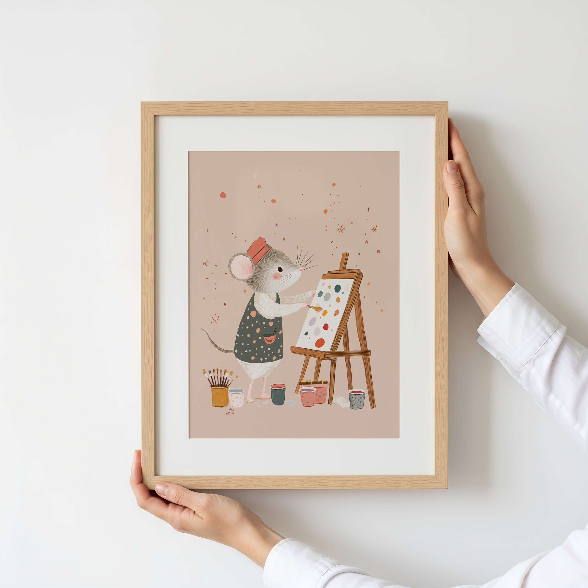 Framed illustration of a mouse painting on an easel held by a person against a white wall.