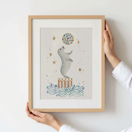 Framed artwork of a seal holding a colorful ball with a person holding the frame.